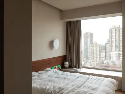 簡約臥室內設有大型窗戶，窗外可見城市景觀，床上鋪著空白的白色毯子，並配有壁燈。