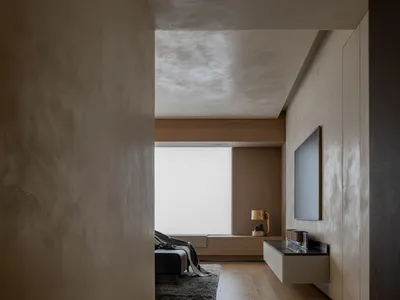 A minimalist bedroom with light wood flooring and furniture, featuring a window with blinds and a carpeted area.