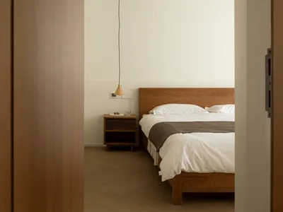 Minimalist bedroom featuring a wooden bed with white bedding and a wall-mounted light fixture against a plain wall.