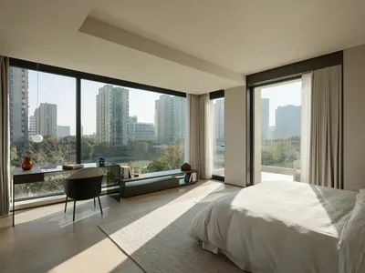 A minimalist bedroom with a large window overlooking city buildings and trees, featuring a bed with neutral linens and a simp
