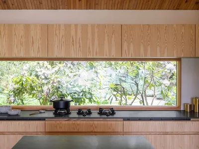 A modern, minimalist kitchen with wooden cabinets and a large window overlooking greenery.