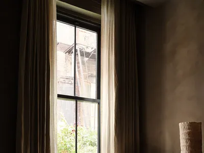 A cozy living room with a white fur-covered armchair, a wooden table, and sheer curtains on a tall window.