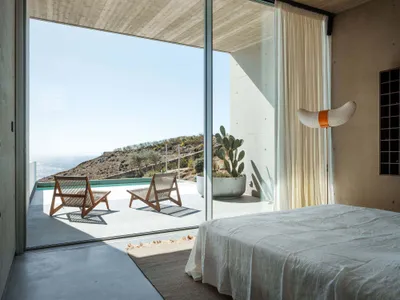 Modern minimalist bedroom featuring white furnishings, concrete walls, and a view of the ocean.