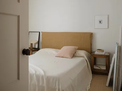 A minimalist bedroom with a bed covered in beige linens, a pink pillow, and a simple headboard.