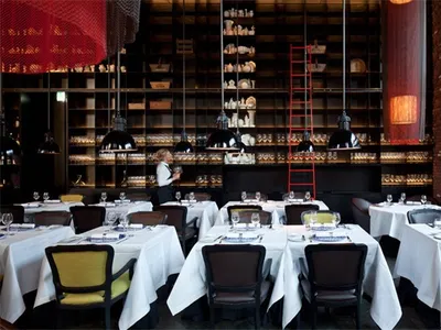 Modern restaurant interior with chairs and tables set with white cloth, multiple pendant lights, and red brick walls.