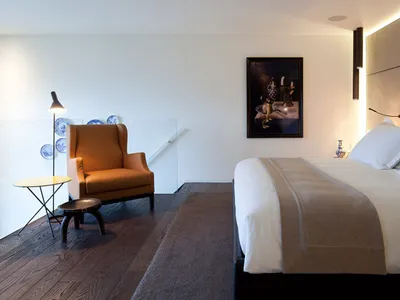 A minimalist bedroom featuring a large bed with a white cloth, an orange armchair, and a wooden floor with a gray rug.
