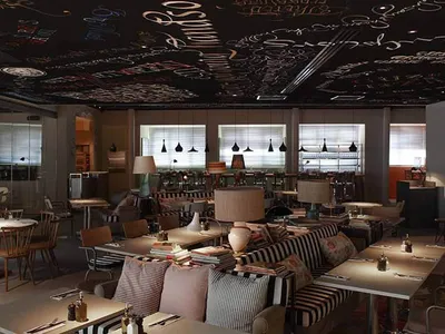 An industrial-style restaurant interior with metal tables, wooden chairs, glass windows, fabric seating, and a chalkboard cei