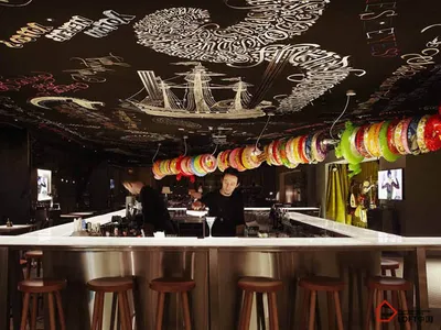 A modern restaurant bar with metal stools, colorful banners, and a chalkboard mural on the ceiling.