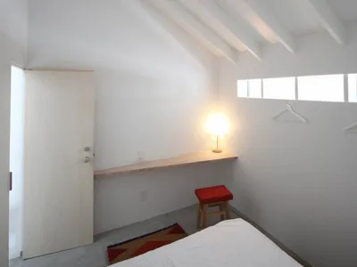 A minimalist bedroom featuring a white bed with a red stool and a lit lamp, under a sloped white ceiling with wooden beams an