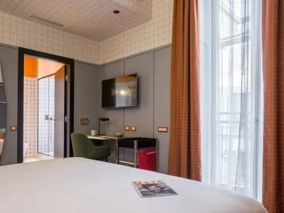 A modern hotel room with a white bed, a bedside table with a red magazine and a green chair, and an orange curtain.
