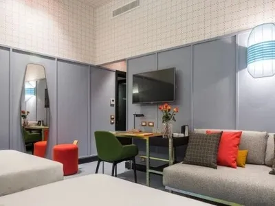A modern hotel room with a grey upholstered sofa, orange cushions, and a television mounted on the wall.