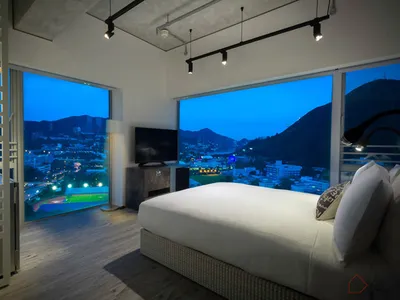 A minimalist bedroom with a large white bed overlooking a cityscape at dusk, featuring sleek, modern furniture and a large wi