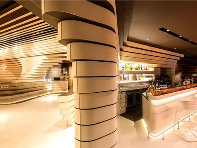 A modern restaurant bar with illuminated wood and metal accents, glass shelves, and seating area.