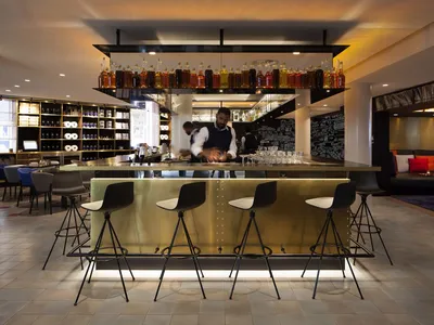 A stylish industrial hotel lobby featuring metallic bar stools, a brass counter, and a variety of alcohol bottles lined up on