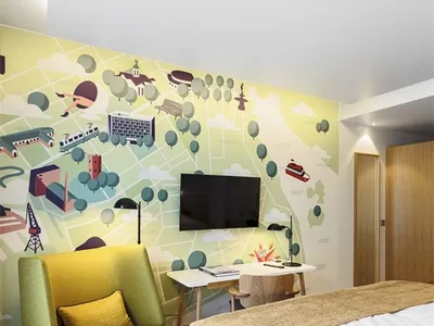 A minimalist bedroom with a green upholstered chair, a bed with a white mattress and beige cover, and a wall mural depicting