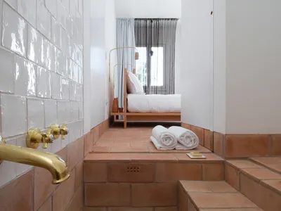 A minimalist bedroom featuring white tiled walls and terracotta tiles on the floor, with a bed and towels.