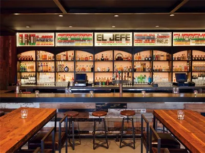 A spacious, modern restaurant bar featuring industrial-style lighting and a wide selection of drinks.