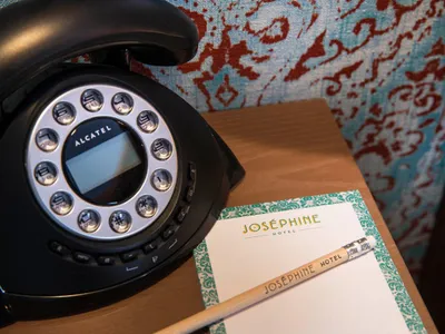 A black rotary telephone and a writing set with a pencil and paper on a wooden desk in a hotel room.