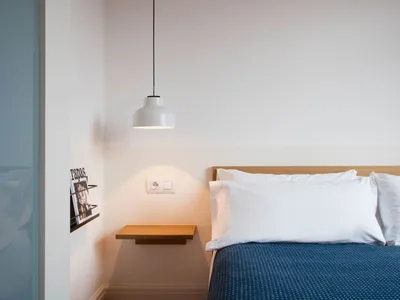 A minimalist bedroom featuring a white bed with a blue blanket and white pillows, under a pendant light and a floating nights