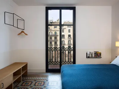 A minimalist bedroom featuring a blue bed with a wooden headboard, white walls, and a large window with a metal railing.