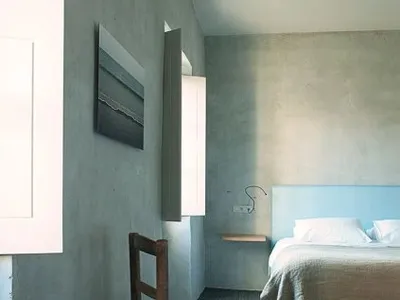 A minimalist bedroom featuring a concrete headboard, white furniture, and a woven chair.