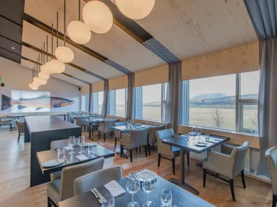 Industrial-style restaurant interior with wooden furnishings and large windows providing natural light.