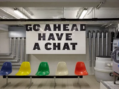 A modern office lounge area with colorful chairs arranged in a row, featuring industrial-style tiles and concrete ceiling.