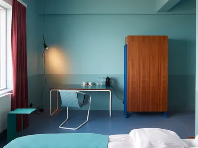 A minimalist bedroom with blue walls, wooden furniture, and a desk with a lamp and coffee mugs.