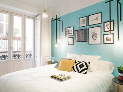 A minimalist bedroom with a light blue accent wall, white furniture, and hanging light bulbs.
