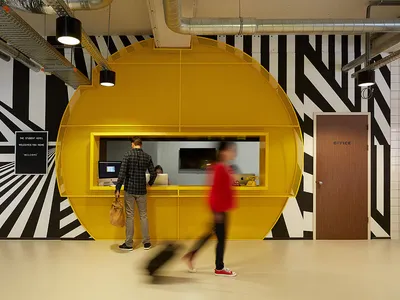 A modern hotel lobby featuring a bright yellow metal reception desk with black and white striped walls.