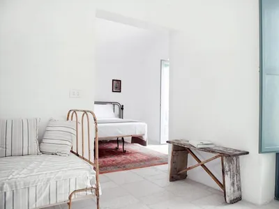 Minimalist bedroom with a white floor, wooden bench, and a bed with white bedding and a single striped cushion.