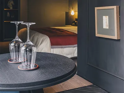A dark-colored wine glass placed upside-down on a wooden table with a black wood floor and a bed with a red blanket in the ba