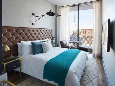 Modern minimalist bedroom featuring a bed with a tufted leather headboard, white bedding, and a teal throw blanket. The room