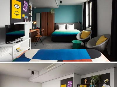 The image depicts a modern minimalist hotel room featuring a bed with a colorful striped bedspread, a desk, a chair, and a te