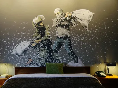 A black and white mural of two individuals engaged in a pillow fight on a bed in a hotel room. The wall features a bird feath