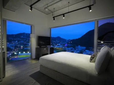 Modern minimalist bedroom with a city view, featuring a large bed, a television, and large windows overlooking the cityscape