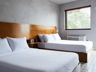 A minimalist bedroom with two double beds, white bedding, and a wooden headboard against a textured wall. The room features a