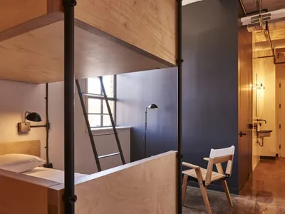 A modern bunk bed with a wooden frame and ladder, featuring a bed with a pillow on the lower bunk and a chair in the foregrou