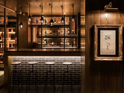 A modern bar interior featuring wooden walls, glass shelves filled with bottles, metal stools, and a brick accent wall.
