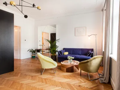 A cozy living room with wooden flooring, a blue sofa, yellow armchairs, and potted plants.