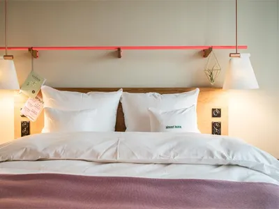 A modern, minimalist bedroom featuring a bed with white pillows and a pink throw blanket, a wooden headboard, and pendant lig
