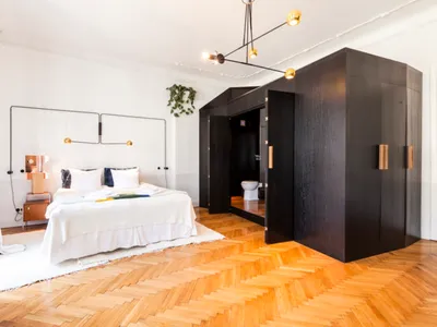 Modern minimalist bedroom with white bed and wooden parquet flooring, featuring a black wardrobe and a plant hanging on the w