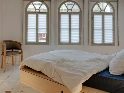 A minimalist bedroom with a white bedframe and blue bedding, featuring arch-shaped windows with white pleated shutters.