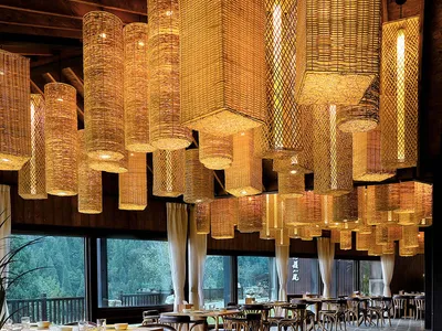 Modern restaurant interior featuring wicker chandeliers and stone flooring, with large windows overlooking a forest.