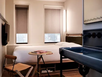Minimalist bedroom featuring a bed with wooden and fabric elements, a small table with a book and glasses, and a metal sink.