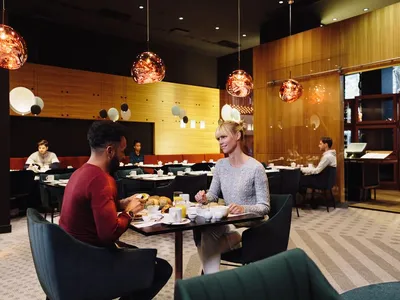Modern restaurant interior with patrons seated at tables, contemporary lighting fixtures, and wooden walls.