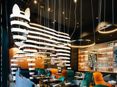 A modern restaurant interior with hanging lights, colorful chairs, and glass-fronted shelves.
