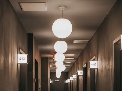 A minimalist hotel corridor with concrete walls, illuminated by spherical pendant lights and numbered room signs.