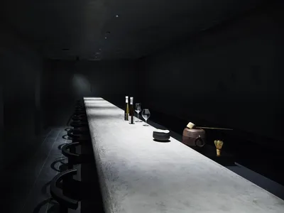 A minimalist restaurant bar with a concrete countertop and black seating on a dark tiled floor.