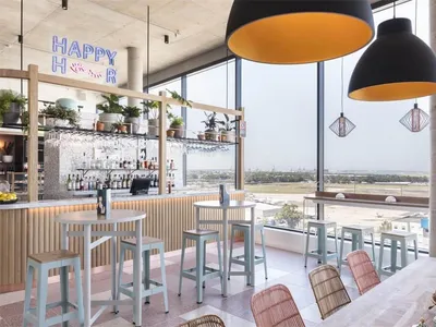 A modern restaurant bar featuring metal composite materials with a Happy Hour sign, high tables, and bar stools overlooking a